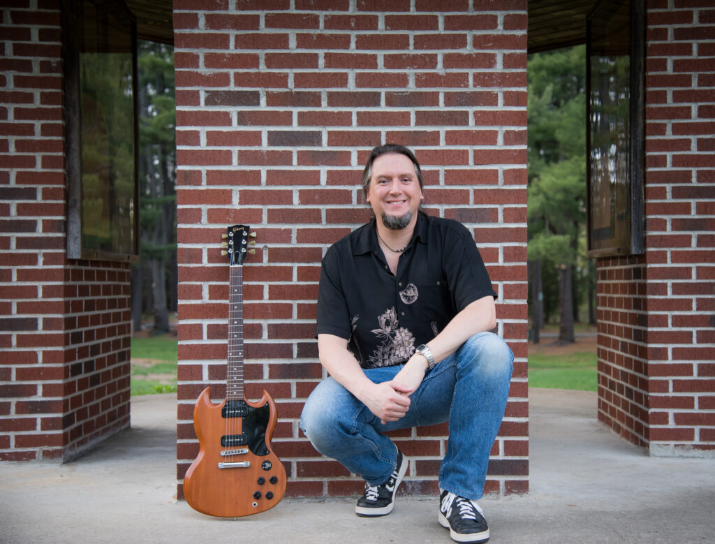 Mike Loce kneeling by brown electric guitar in front of a brick wall